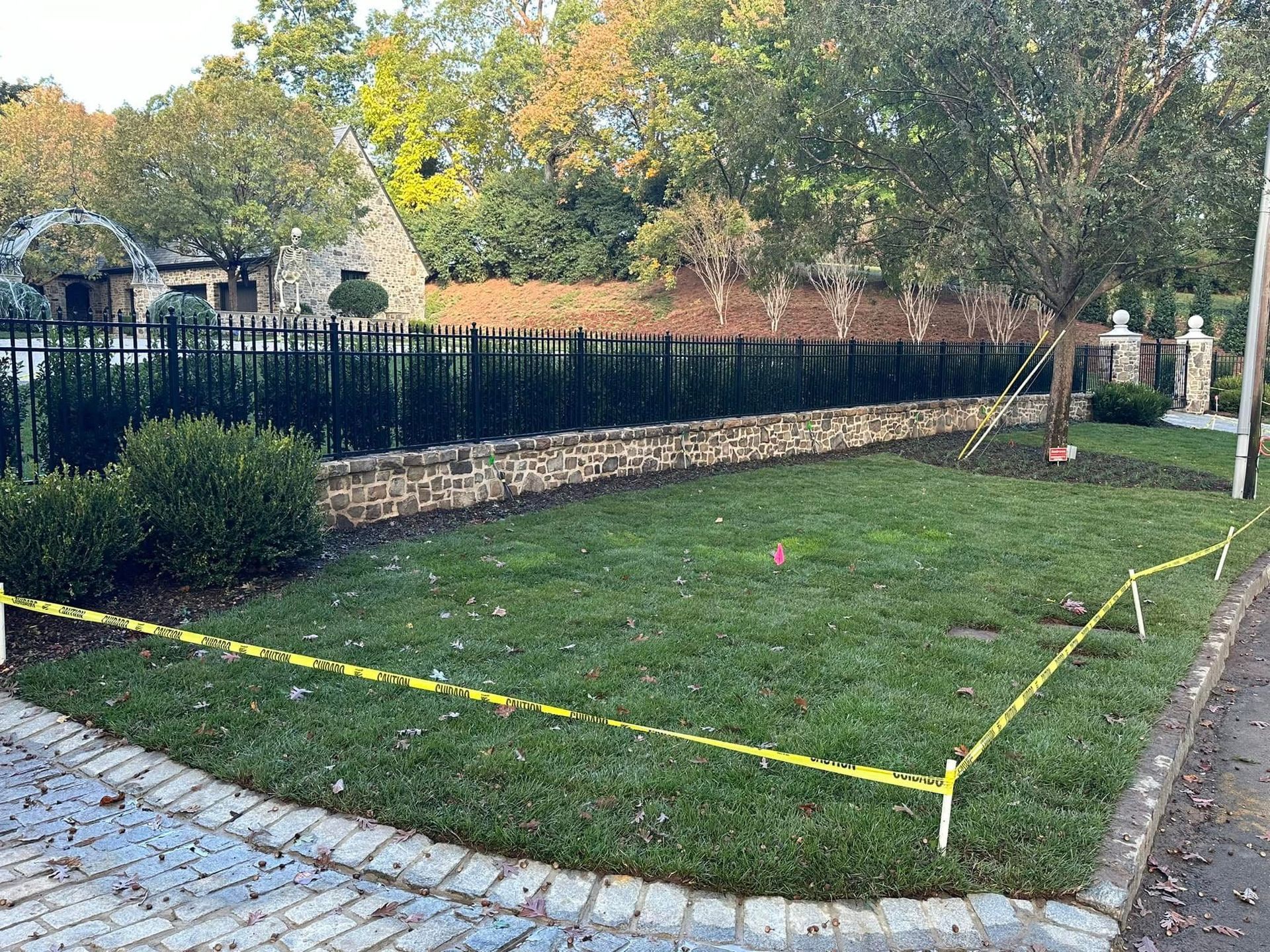 Marked grassy yard beside a stone walkway and black fence, with yellow caution tape outlining a rectangular area.