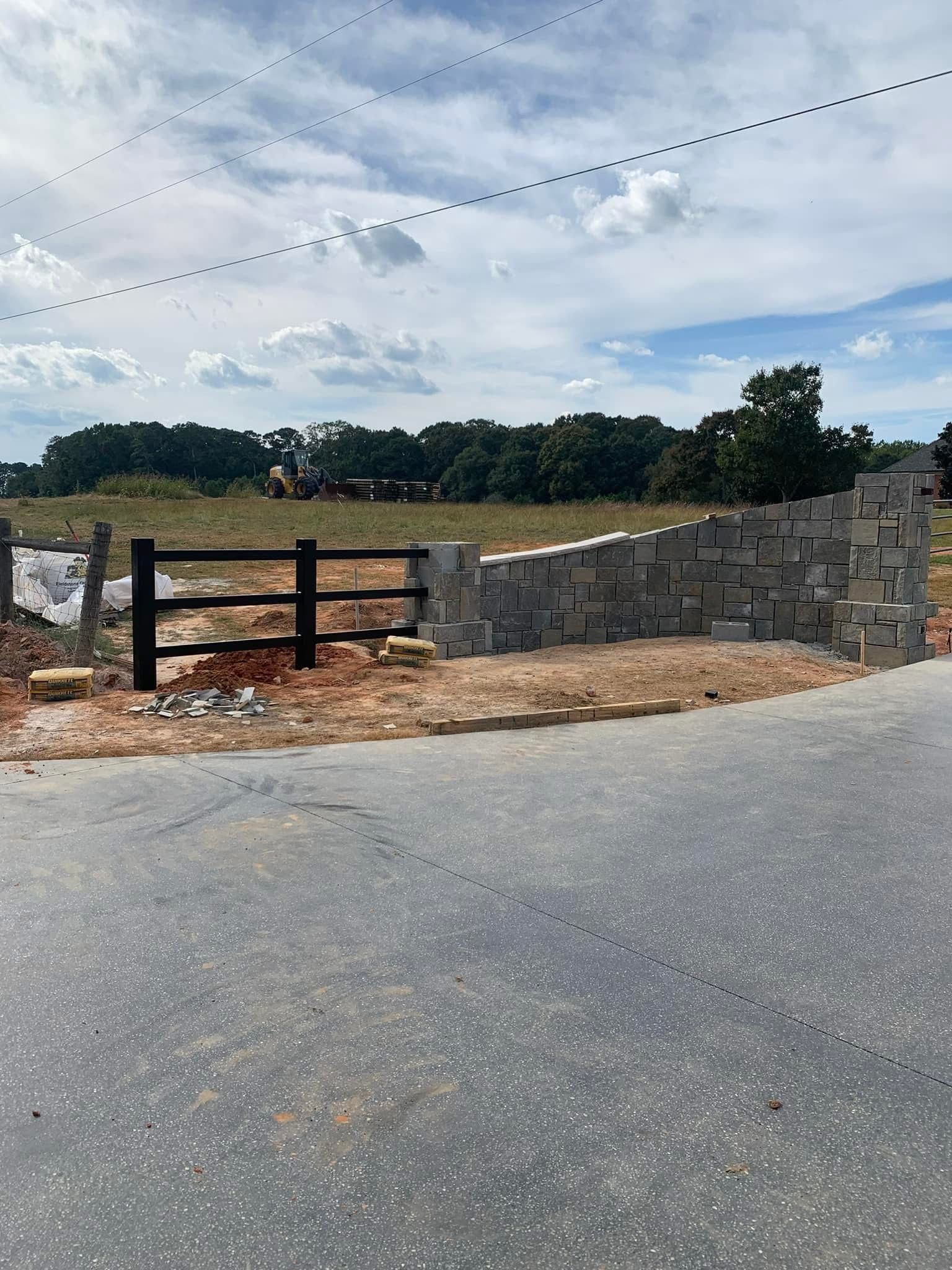 Stone wall and wooden gate along a roadside beside an open field under a cloudy sky.