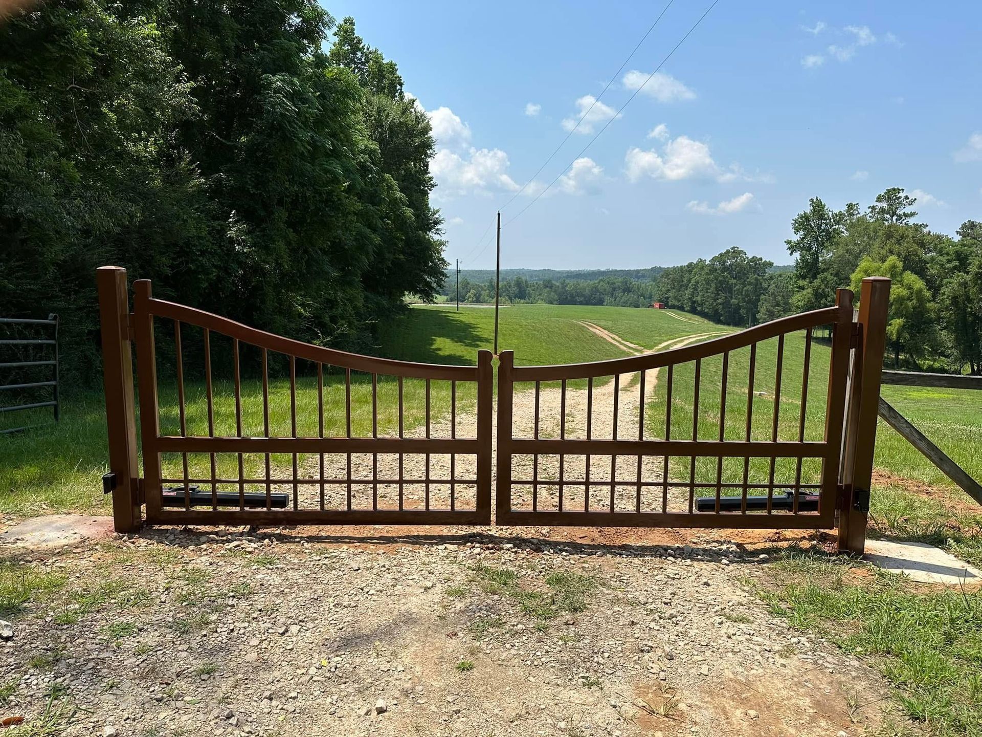 Wooden ranch gate closed across a gravel driveway, opening to a green field and tree-lined country road.