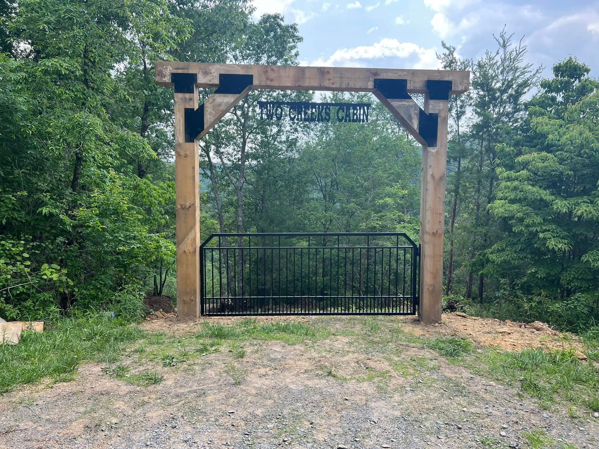 Wooden garden gate with black metal gate beneath a rustic arbor, surrounded by trees and gravel path