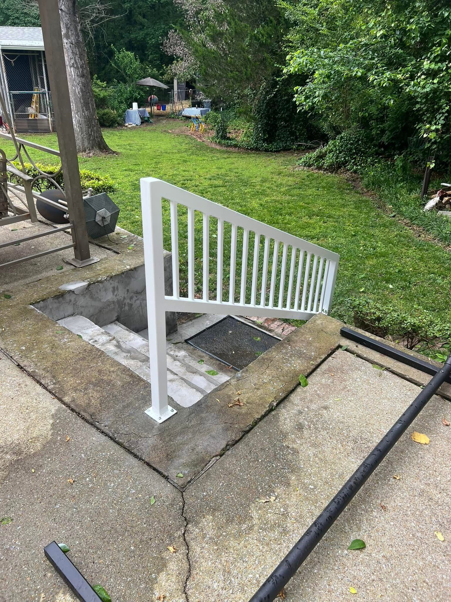 Basement stairwell entrance with white railing set into a concrete patio beside a grassy yard.