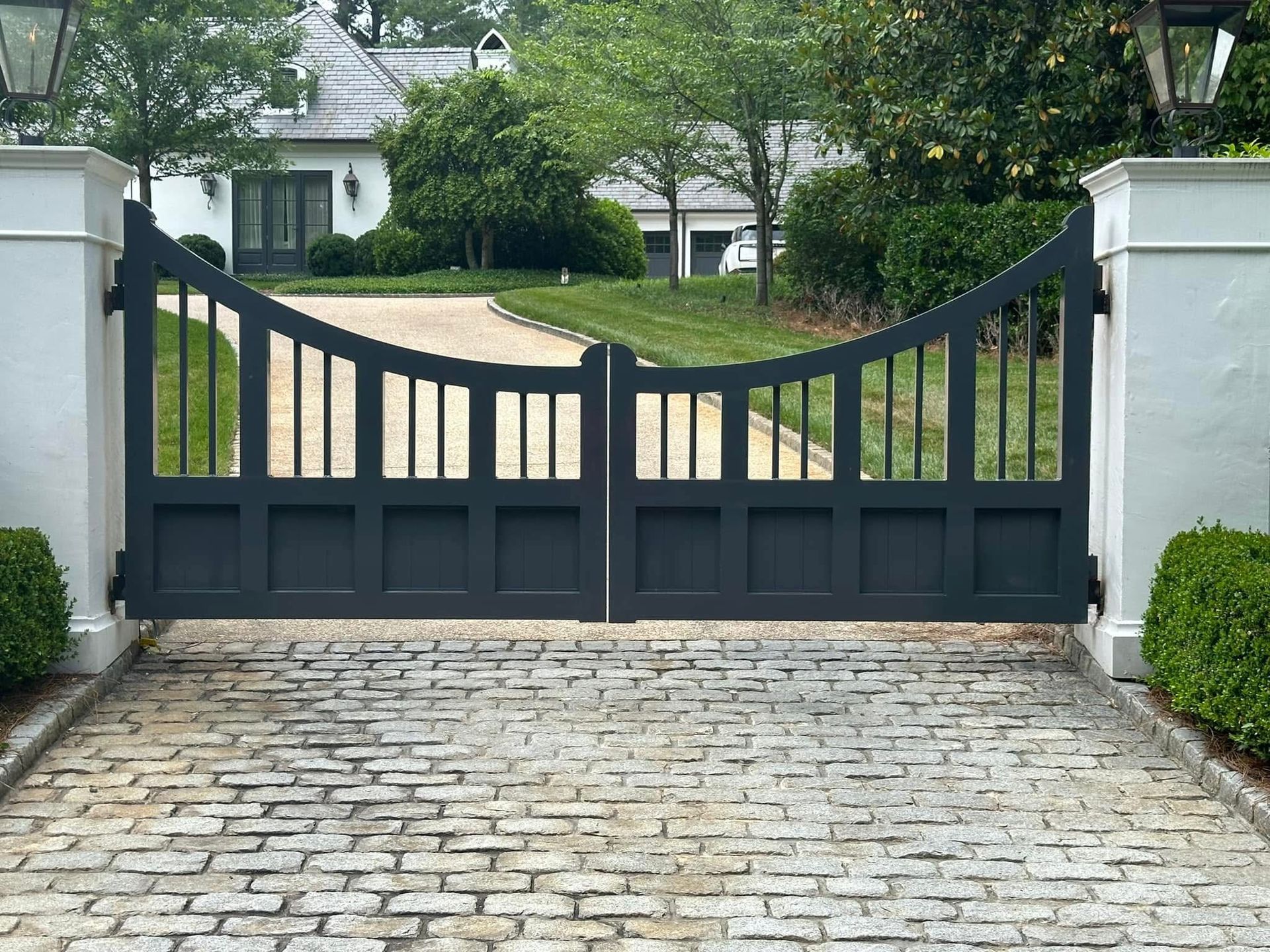 Dark double gate at the end of a cobblestone driveway with white pillars and greenery