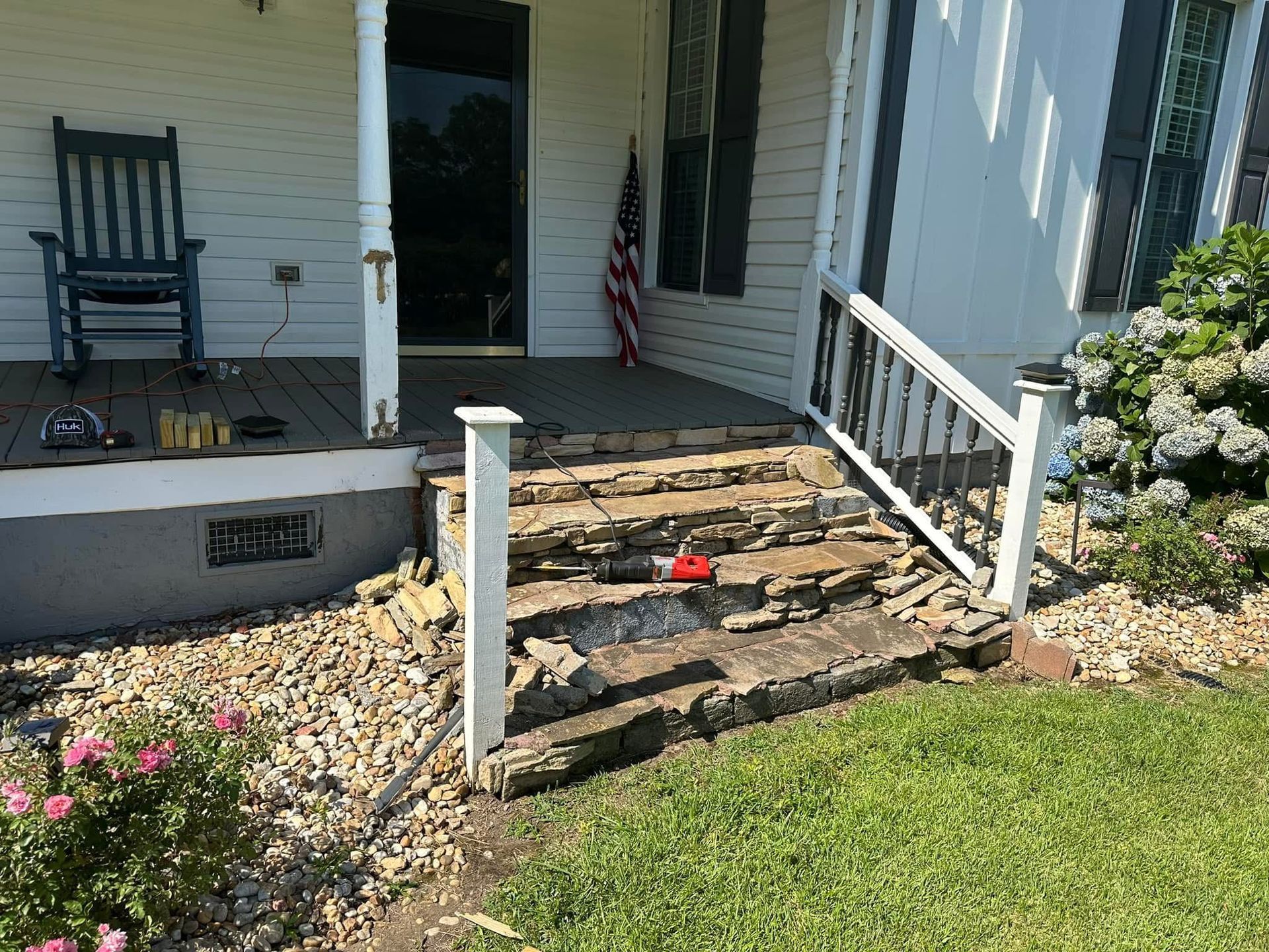 Front porch steps with white railings, a black chair, and a small flower bed beside a lawn.