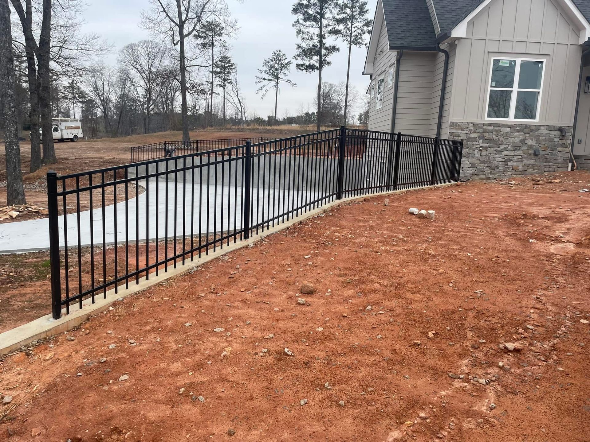 Black metal railing beside a curving concrete path on a red dirt slope near a stone house