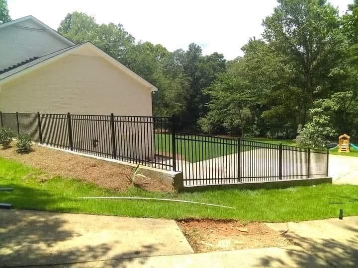 Backyard patio with black metal fence, sloped lawn, and beige house beside trees and driveway
