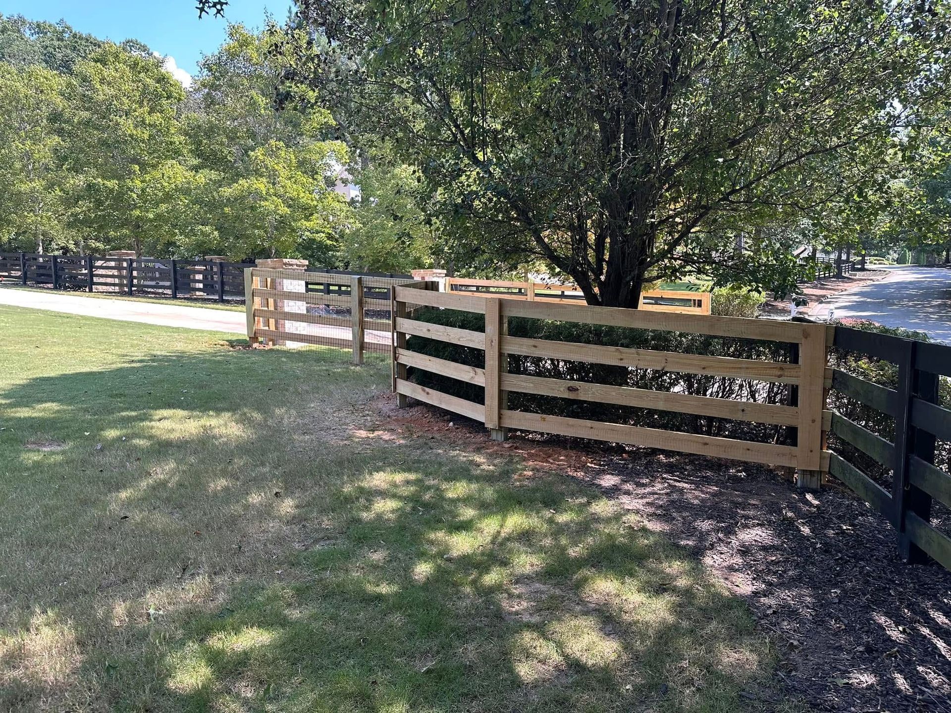 Wooden fence along a grassy path beside trees, with sunlight and shadows in a wooded park-like area.