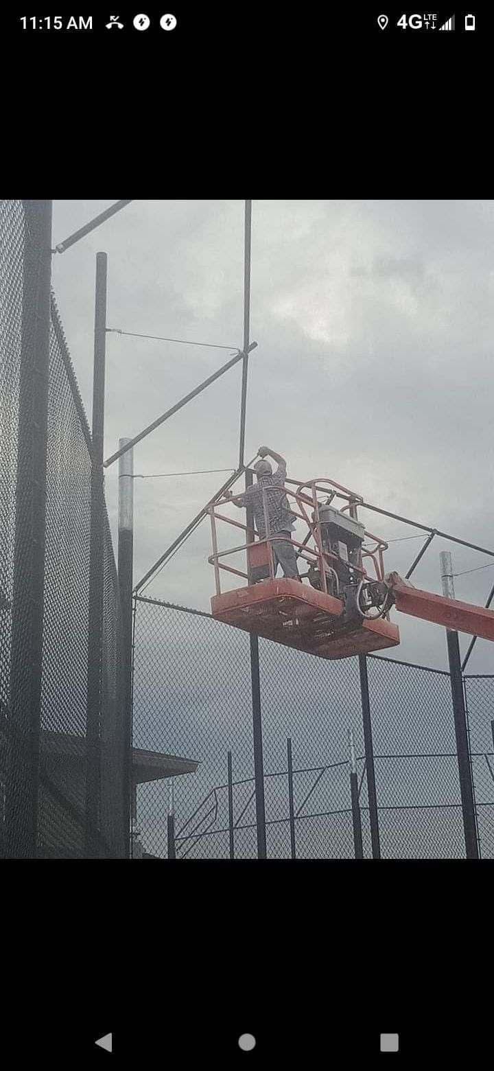 Person on an orange cherry picker working near a tall chain-link fence under a cloudy sky