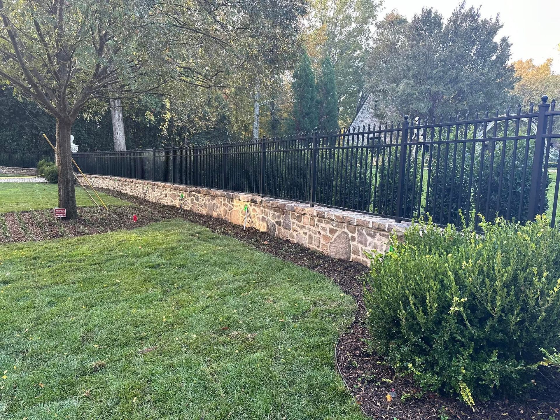 Stone retaining wall beside black metal fence along a grassy yard with shrubs and trees