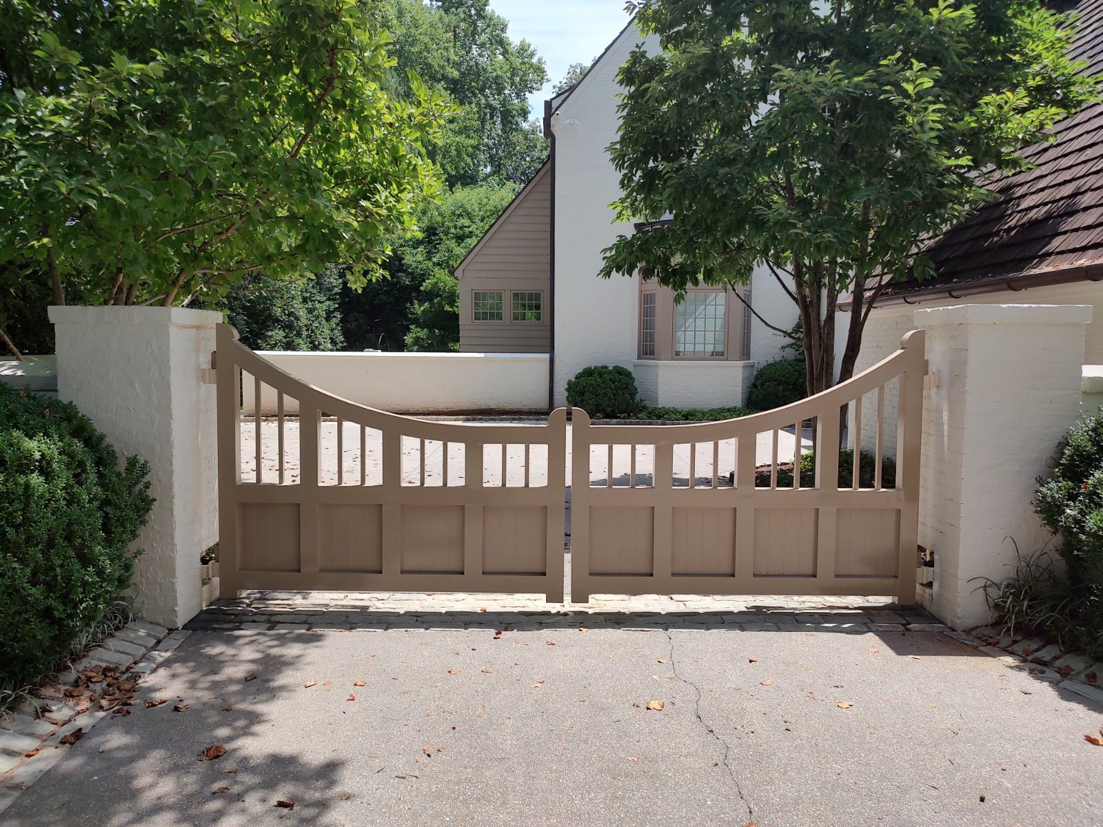 Beige wooden driveway gate with curved top, set in a sunny residential entrance with trees and white walls
