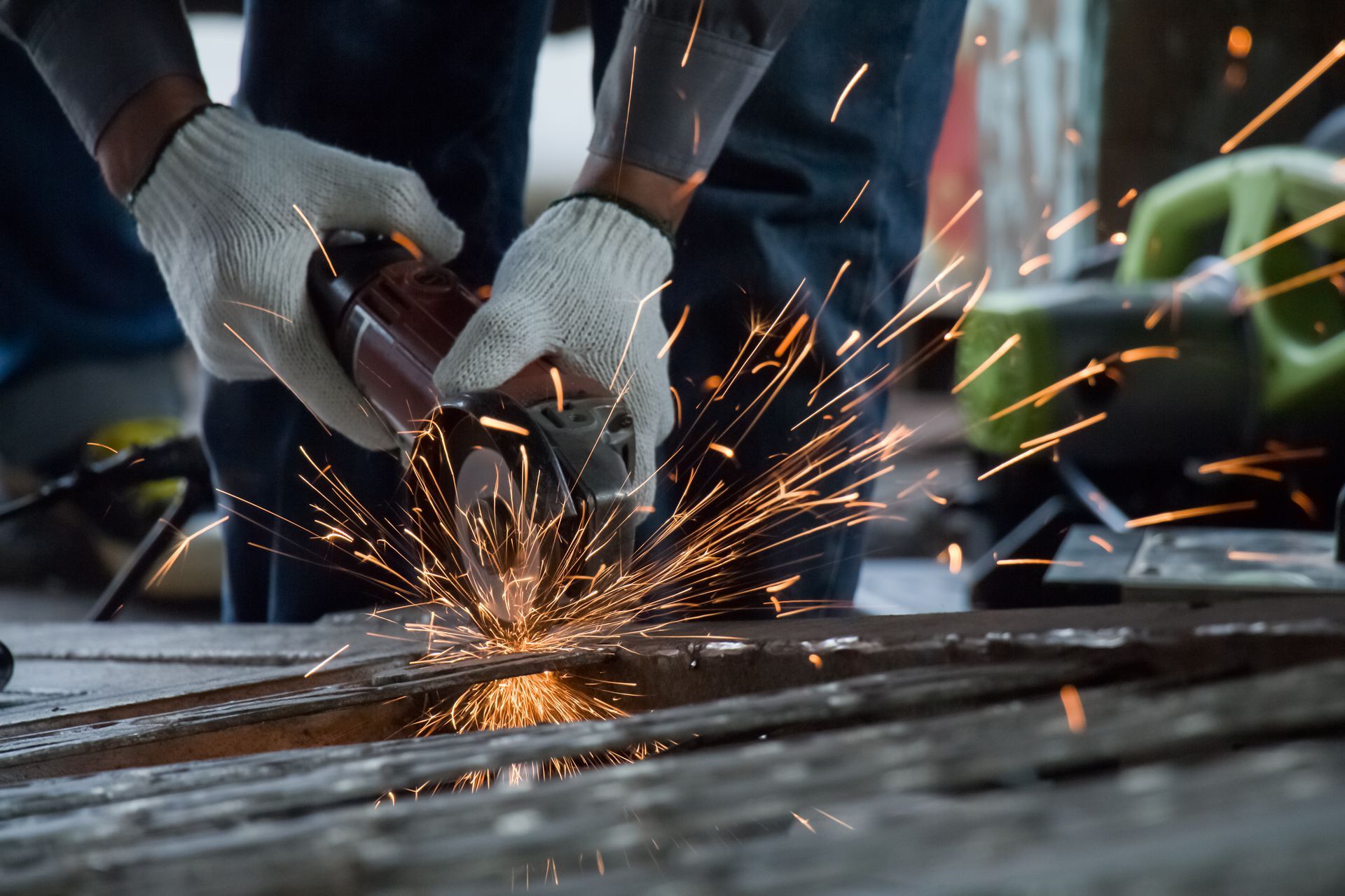 Gloved hands cutting metal with an angle grinder, sending bright sparks across a workshop surface