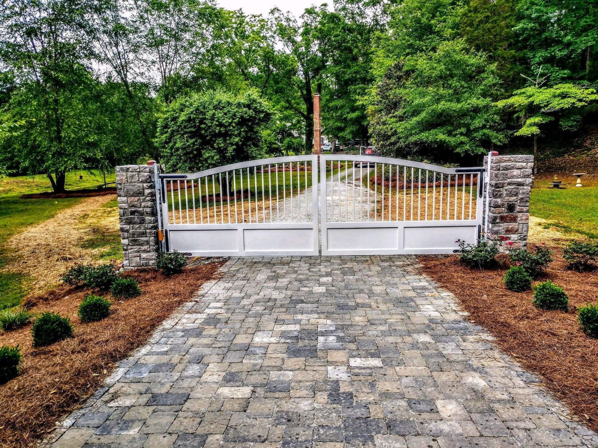 White metal gate at the end of a cobblestone driveway, flanked by stone pillars and trees.