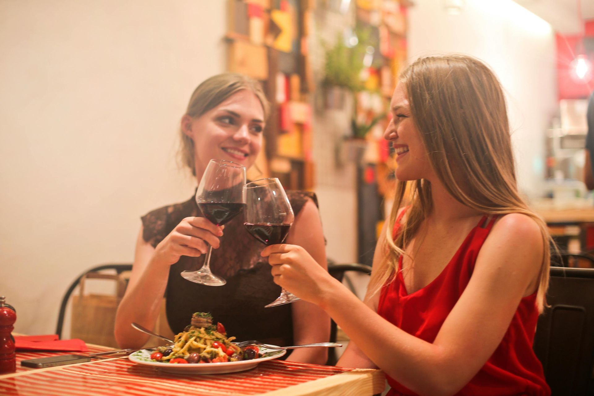 Two women are sitting at a table toasting with wine glasses.