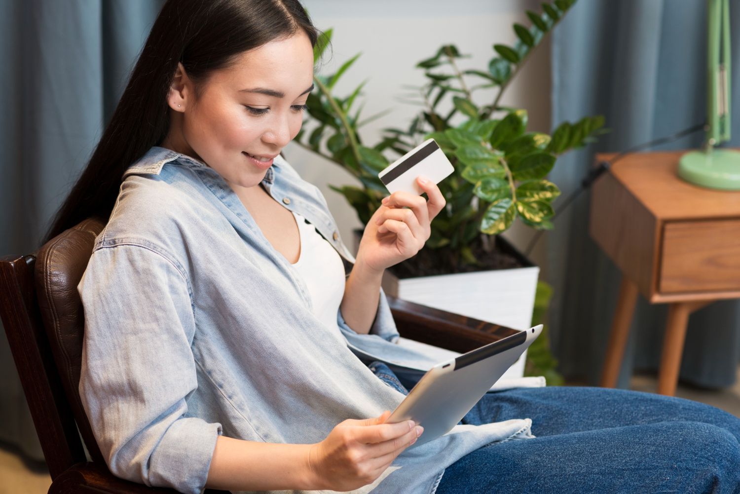 A woman is sitting in a chair holding a credit card and a tablet.