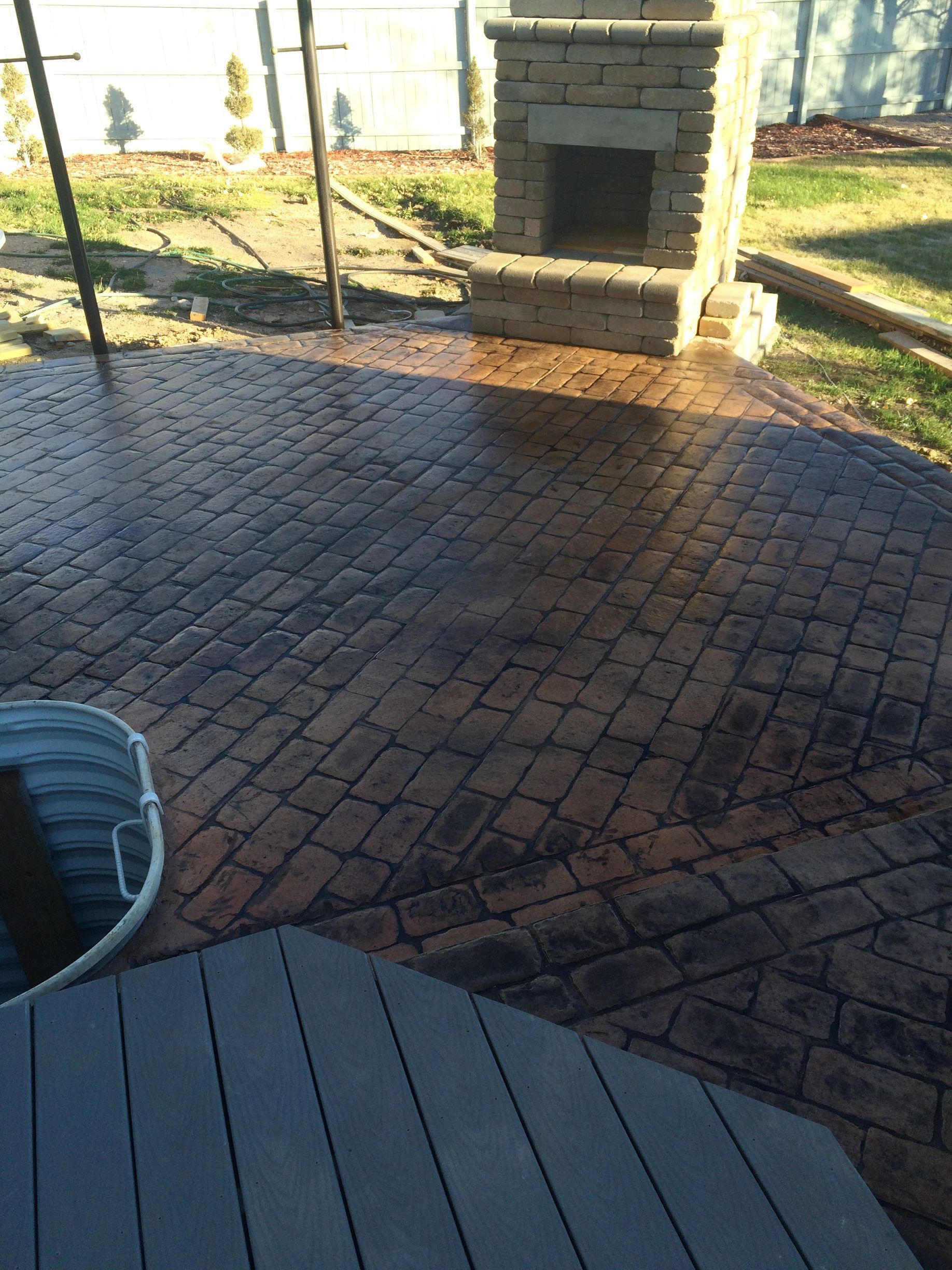 Brick-patterned stamped concrete patio with an outdoor fireplace in a yard; a table and a bucket are also visible.