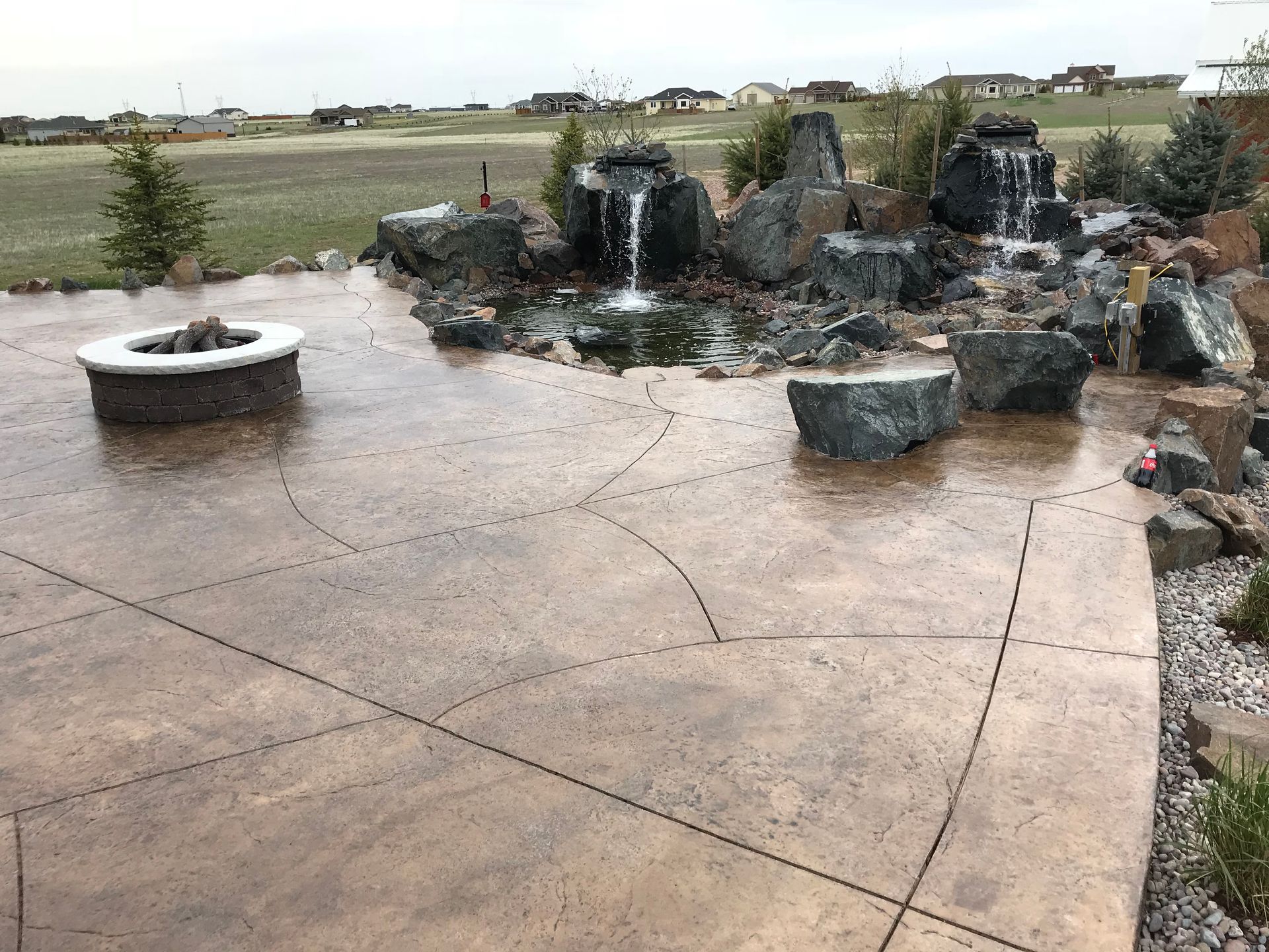 Patio with rock waterfall feature, fire pit, and textured concrete flooring in an outdoor setting.