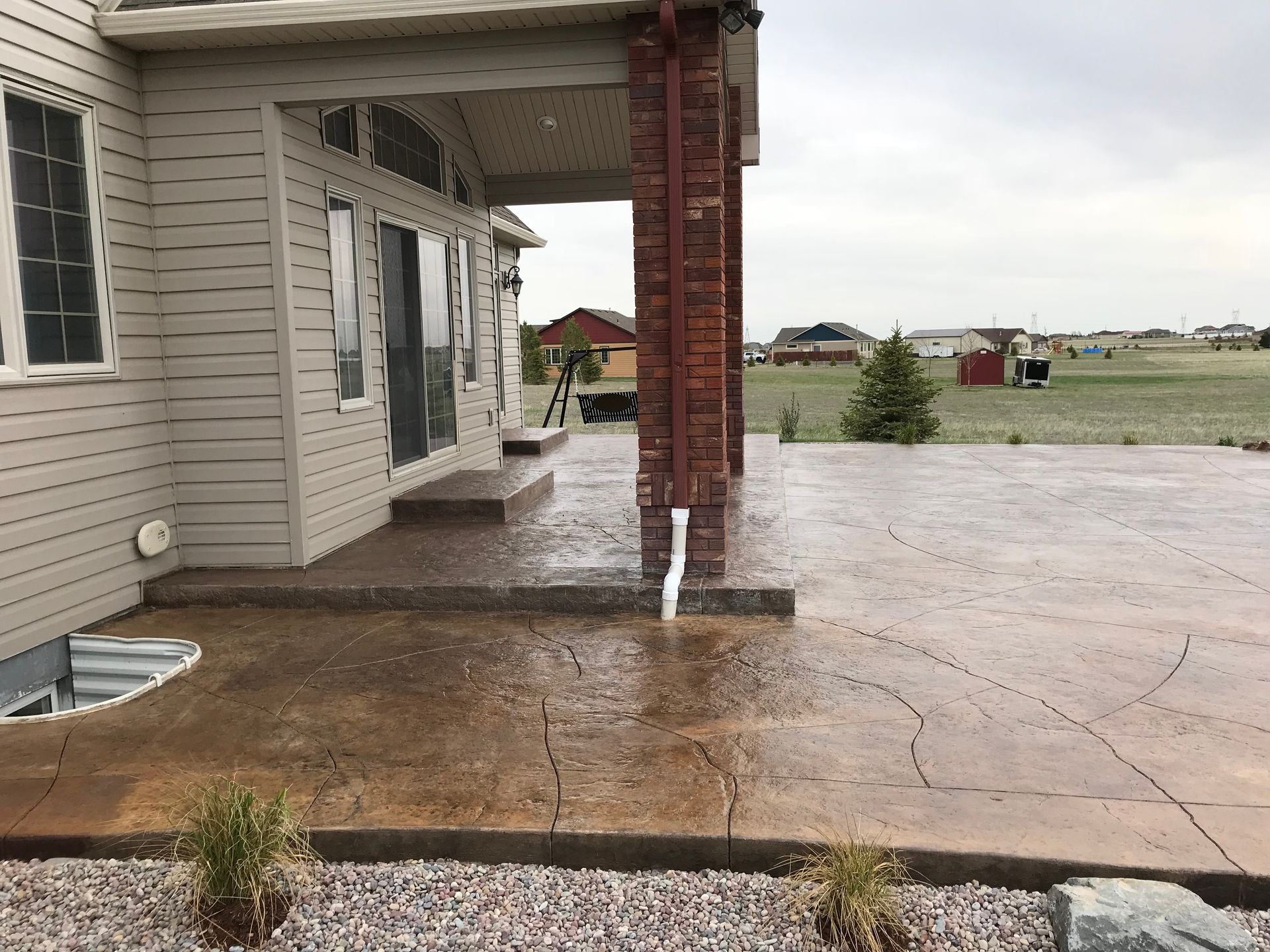 Back patio with textured concrete, a brick column, and a grassy yard under a cloudy sky.
