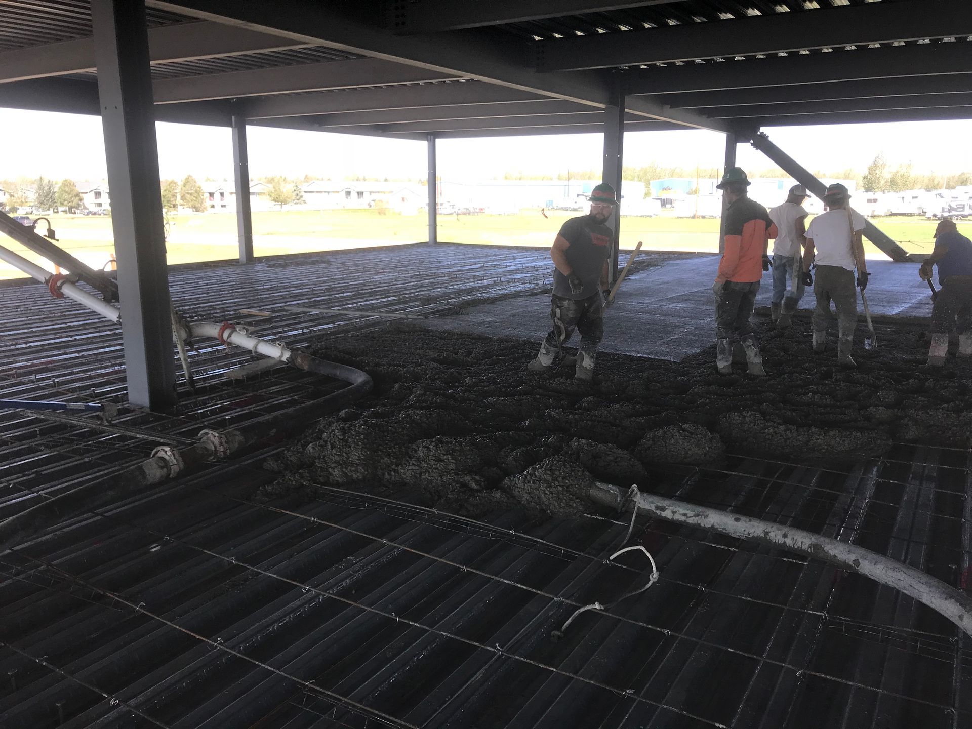 Construction workers pouring concrete on rebar, under a steel-framed structure.