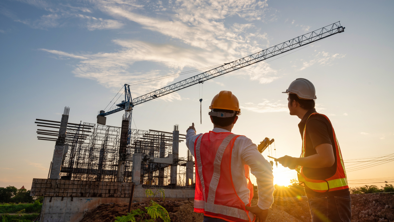 Two construction workers in hard hats and safety vests look toward a building site with a crane at sunset.