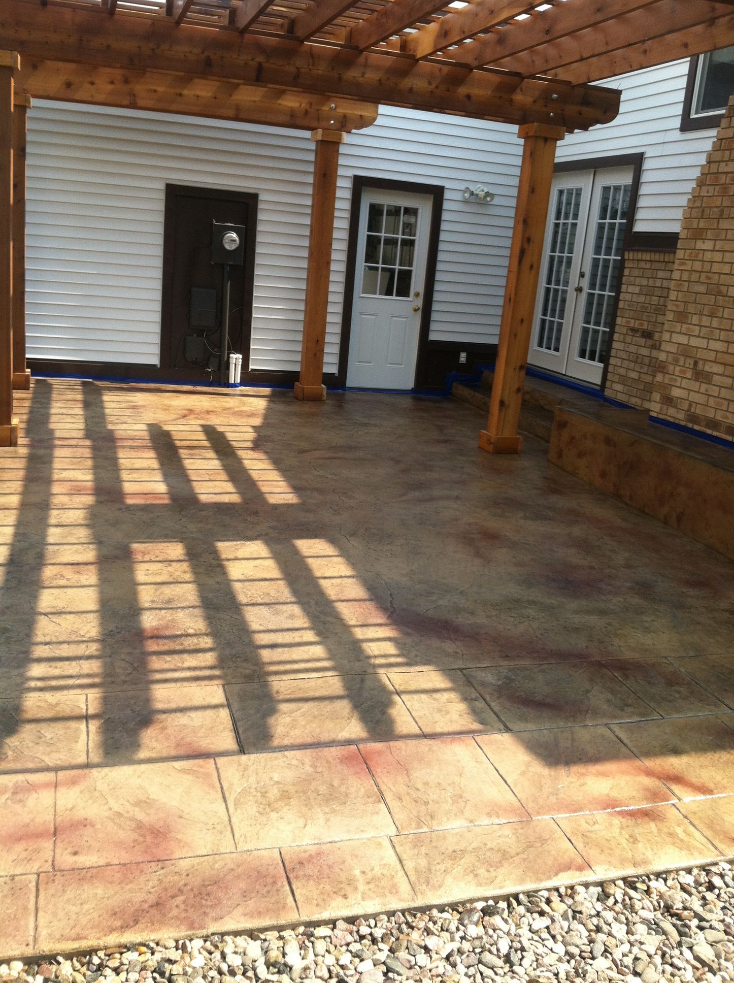 Pergola over a concrete patio with brick border, next to a white house with multiple doors.