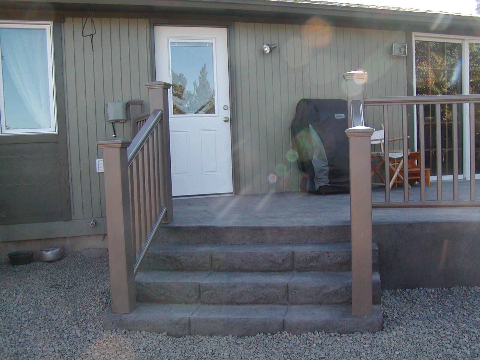 Exterior view of a house with steps leading to a door, a small deck with railing, and gravel ground.