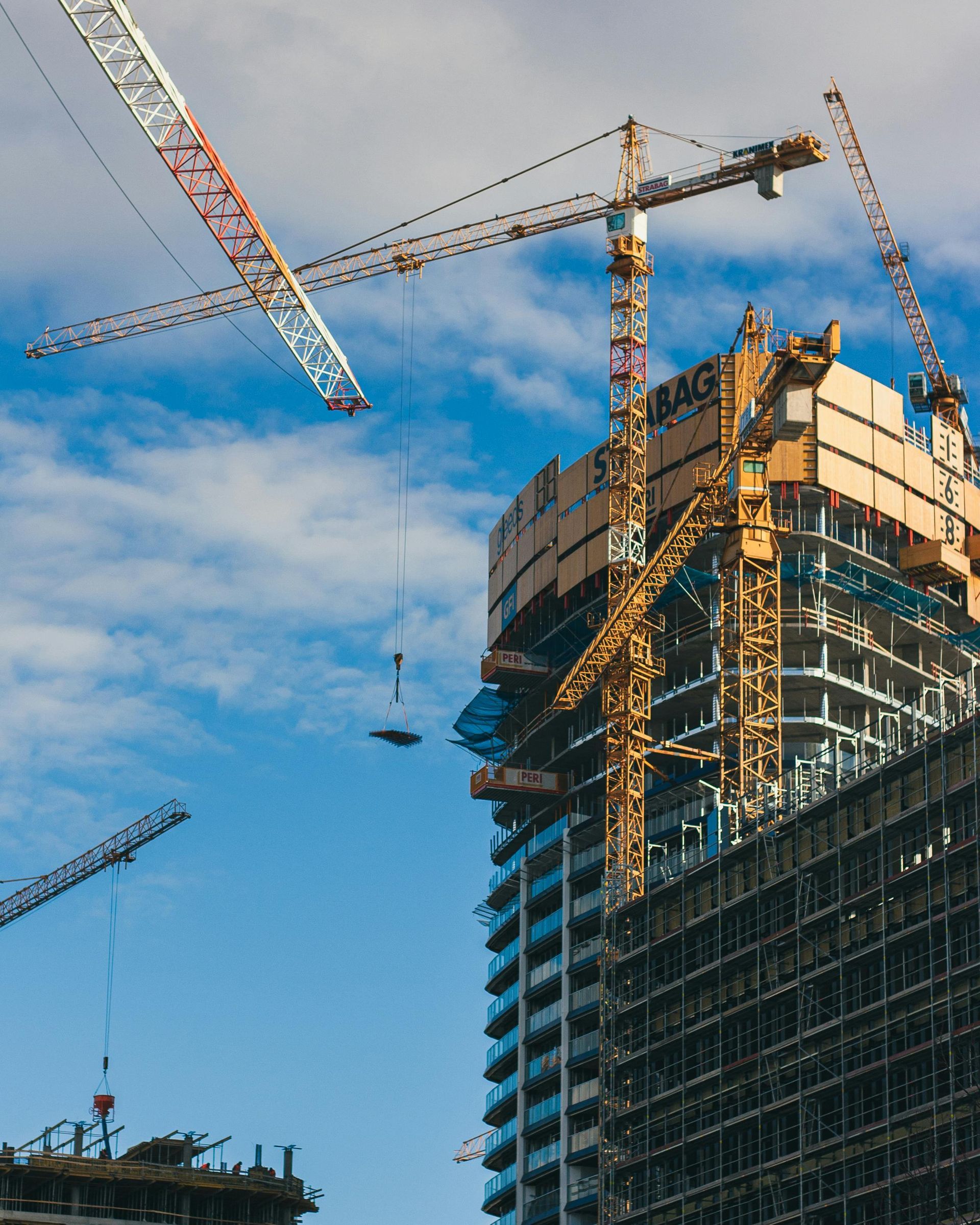 Construction site with several cranes lifting materials to a tall building under construction against a blue sky.