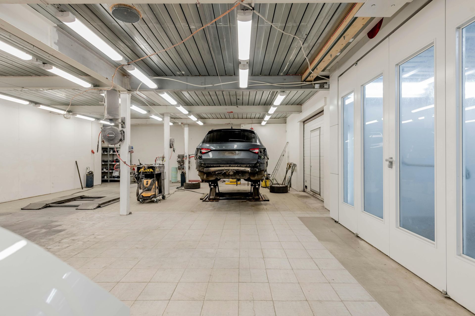 Car on a lift in a bright auto body shop with spray paint booth, tools, and lights.