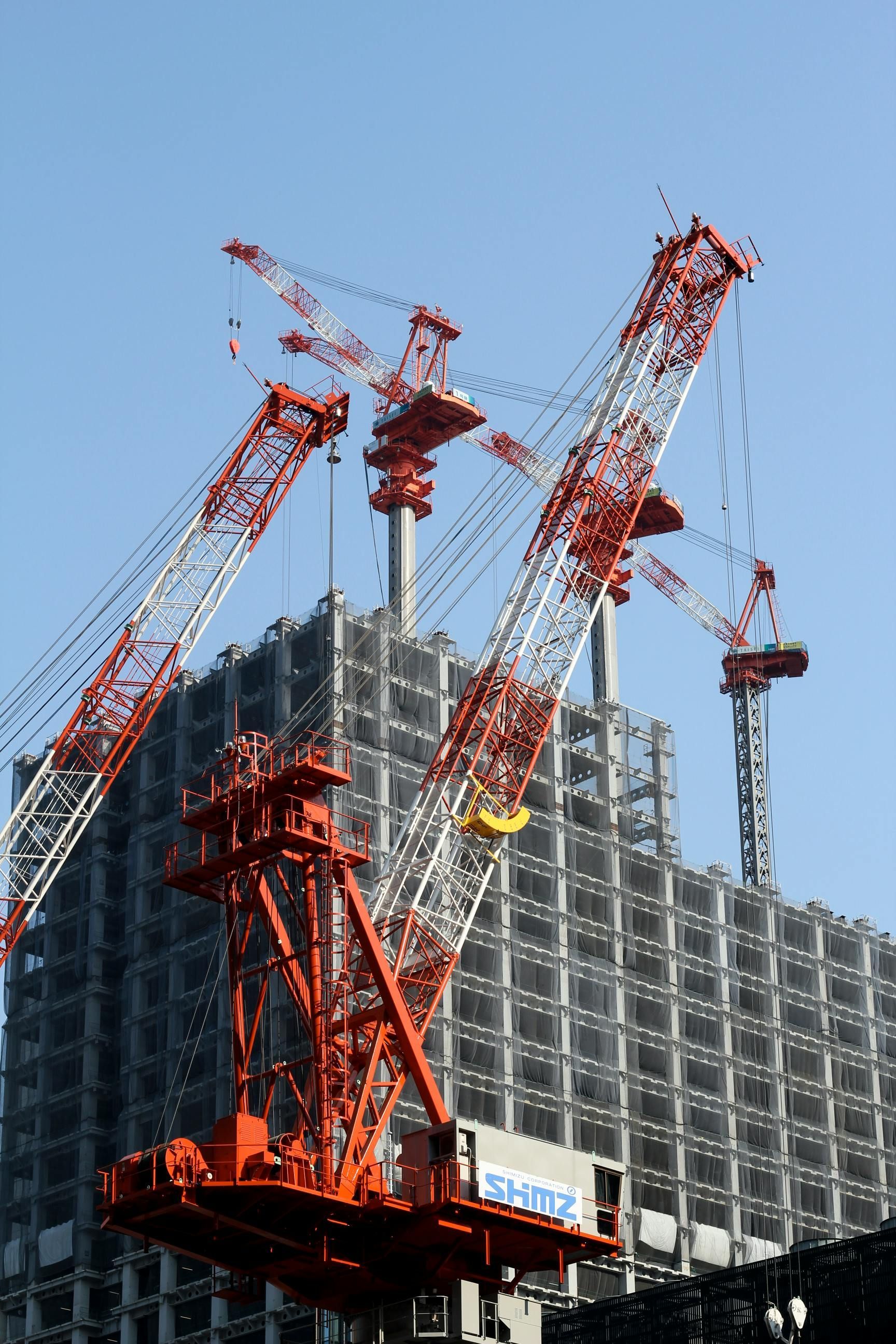 Construction cranes at work on a high-rise building with a steel frame, against a clear blue sky.