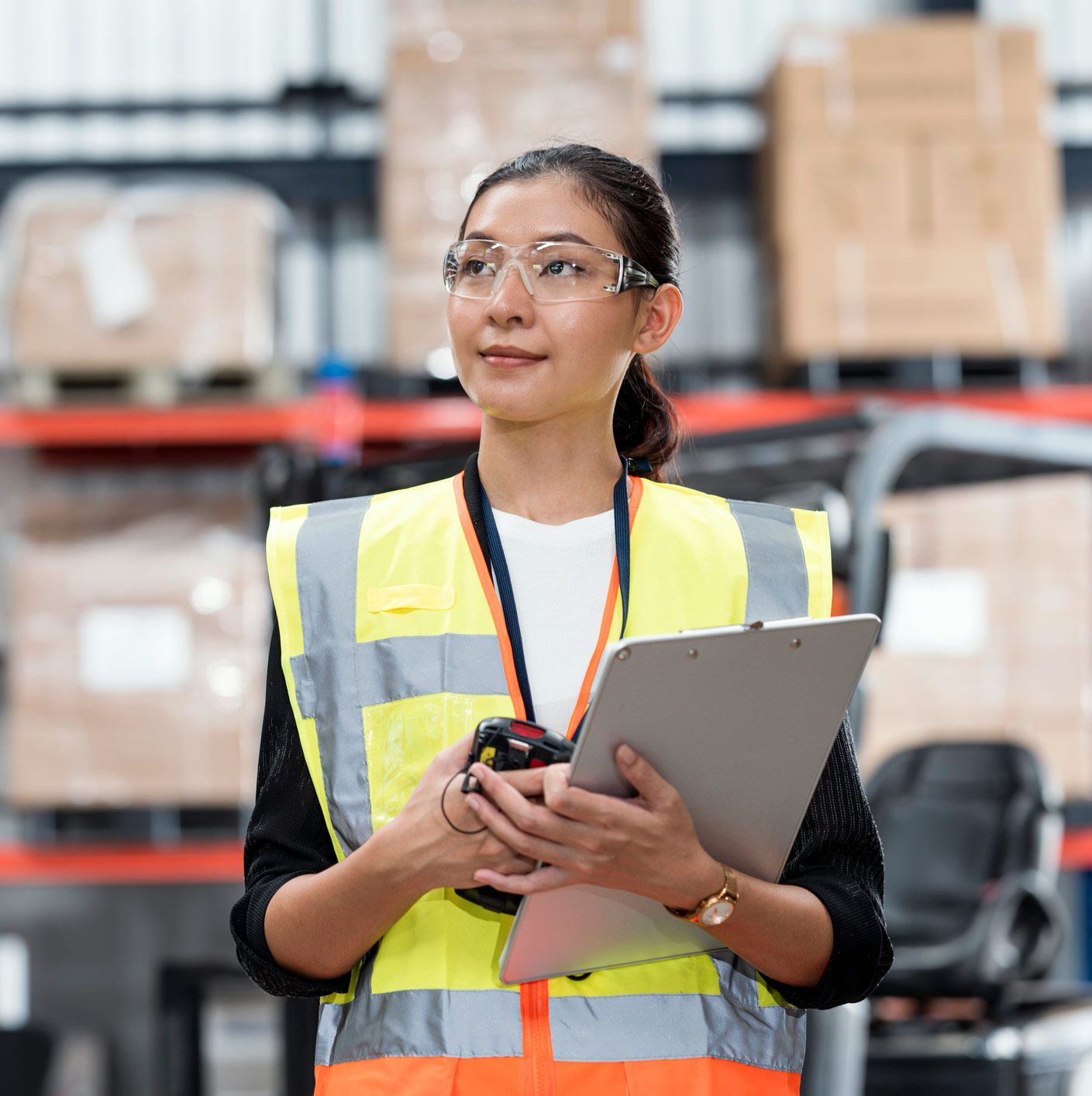 A woman in a yellow vest is holding a clipboard in a warehouse.