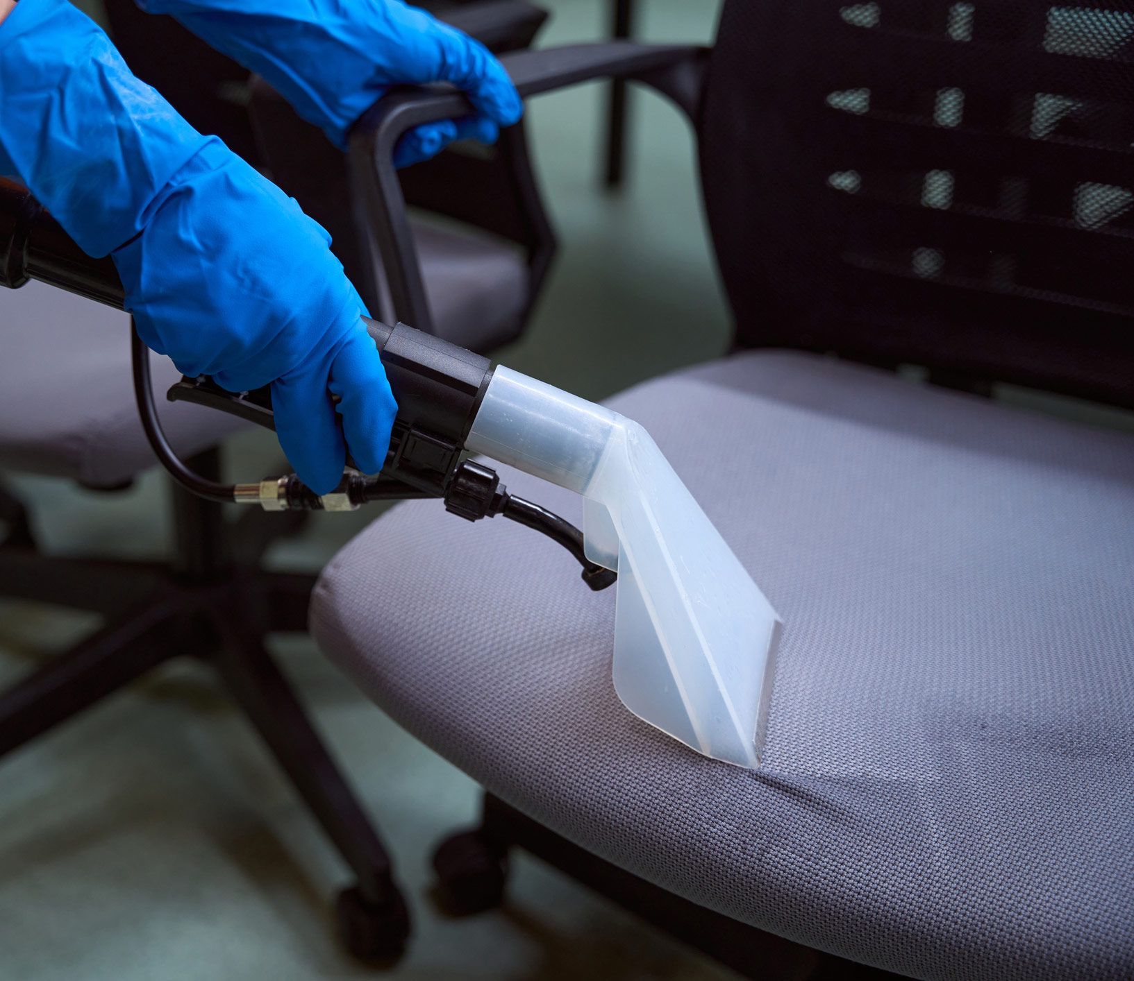 A person wearing blue gloves is cleaning an office chair with a vacuum cleaner.