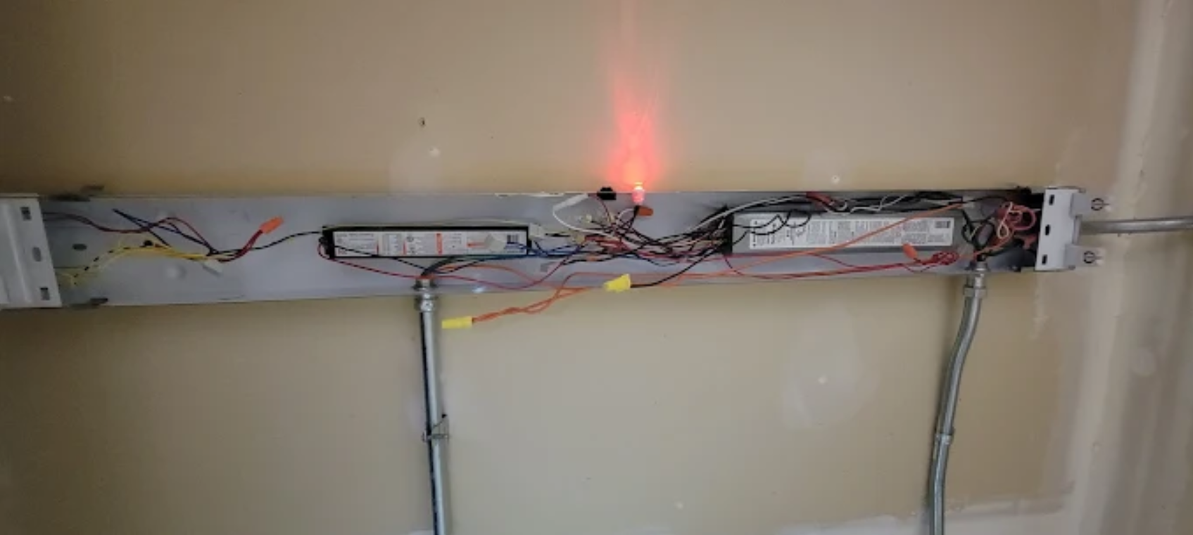 Exposed electrical wiring in a wall opening with a glowing red indicator light and two metal conduit pipes below.