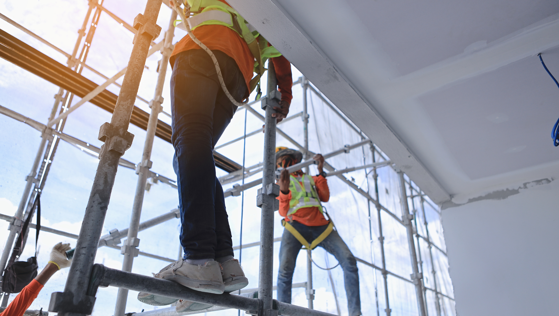 Construction workers on scaffolding, wearing safety vests and harnesses, working near a white ceiling.
