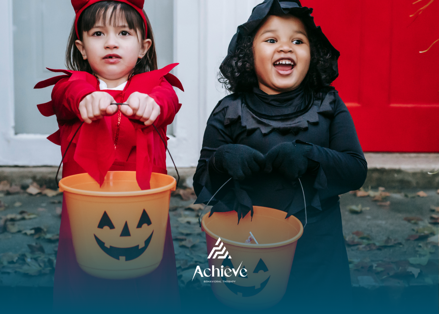 Two children in red and black Halloween costumes hold pumpkin treat buckets outdoors.