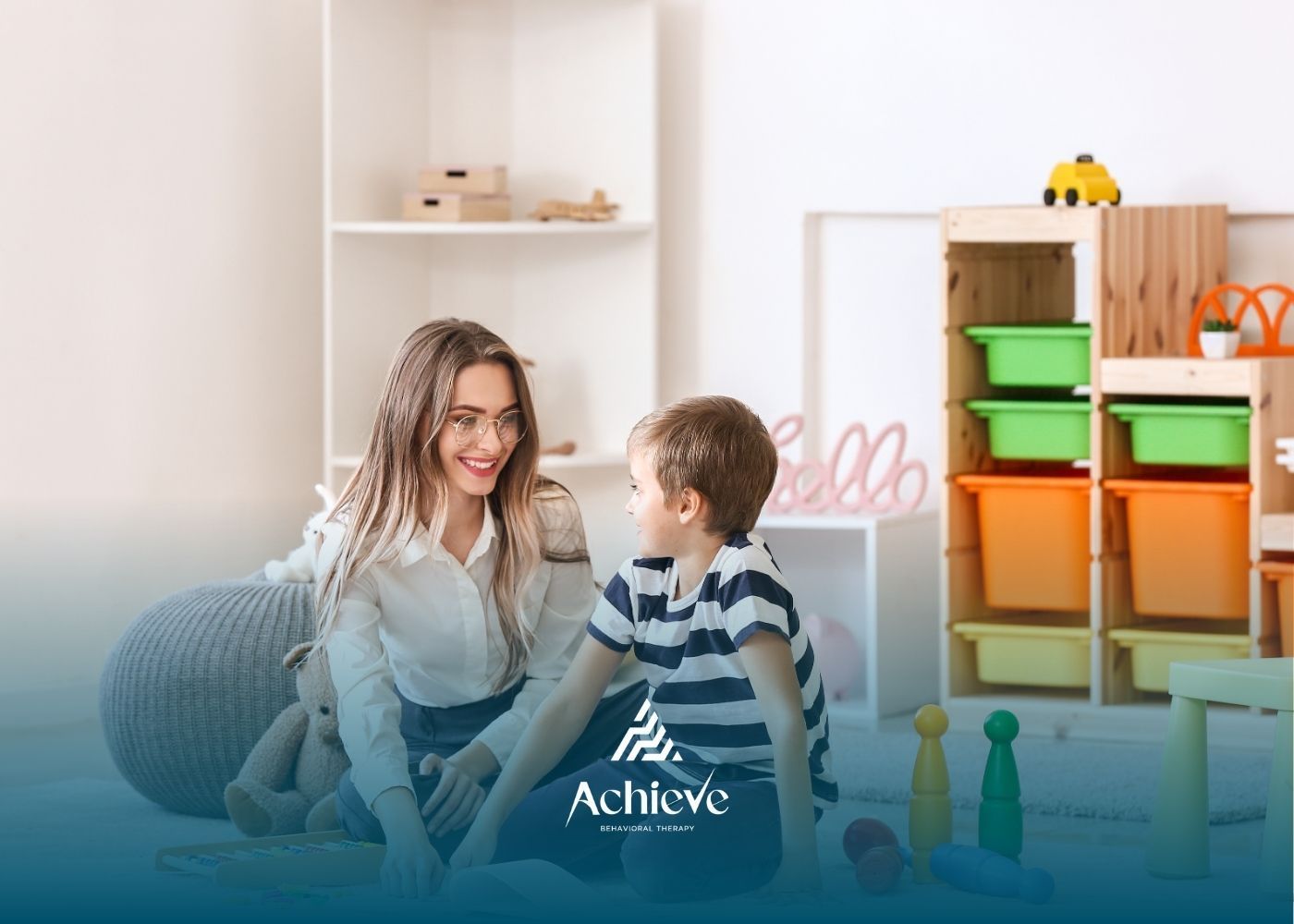 Woman smiles at a child in a playroom. They are seated on the floor. Shelves and toys in the background.