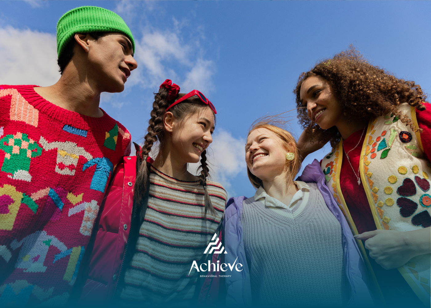 Four friends smiling outdoors beneath a blue sky, wearing colorful winter clothes