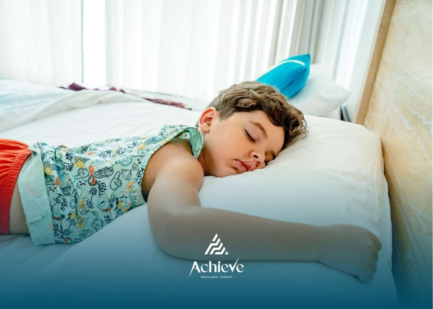 Boy sleeping on a white pillow, wearing a patterned top and red shorts. Bright bedroom setting.