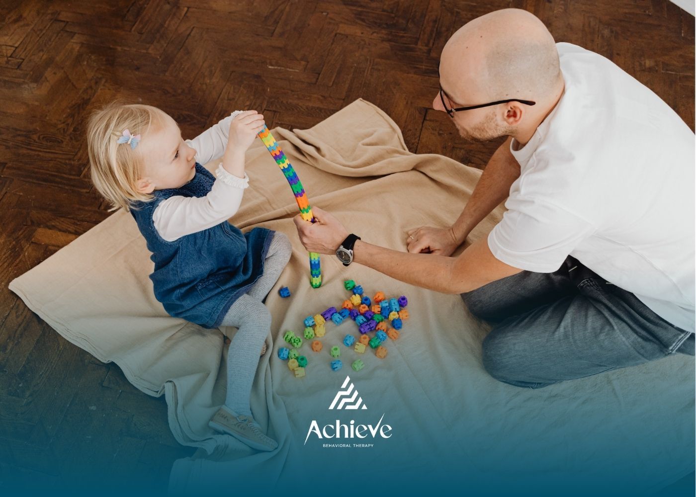 A person and a young child sit on a blanket, building a vertical tower with small, colorful, interlocking plastic pieces.