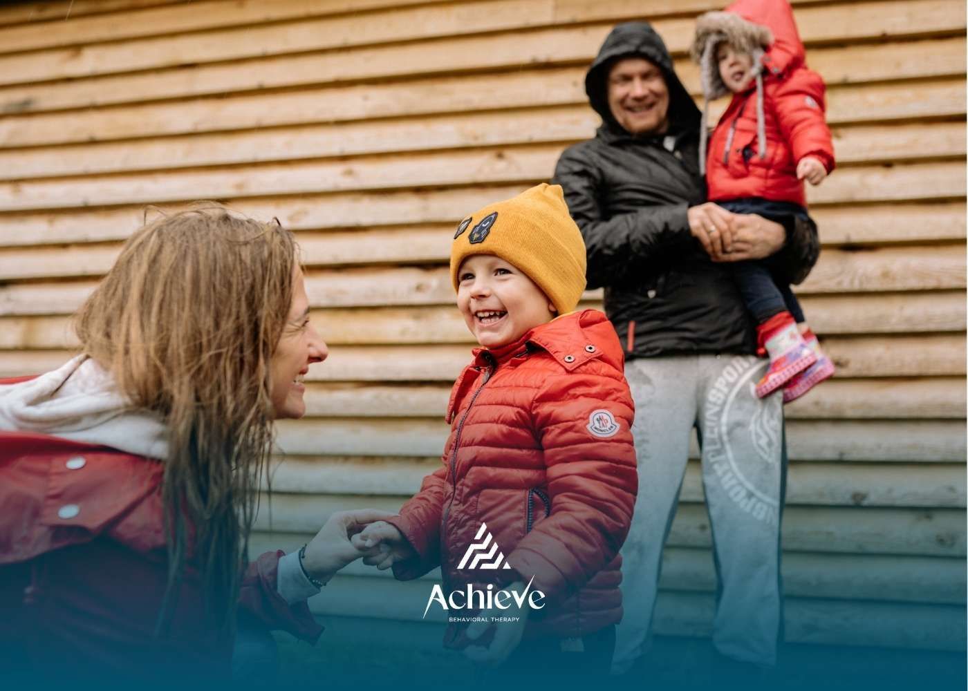 Family of four smiles by wooden wall; father holds a child.
