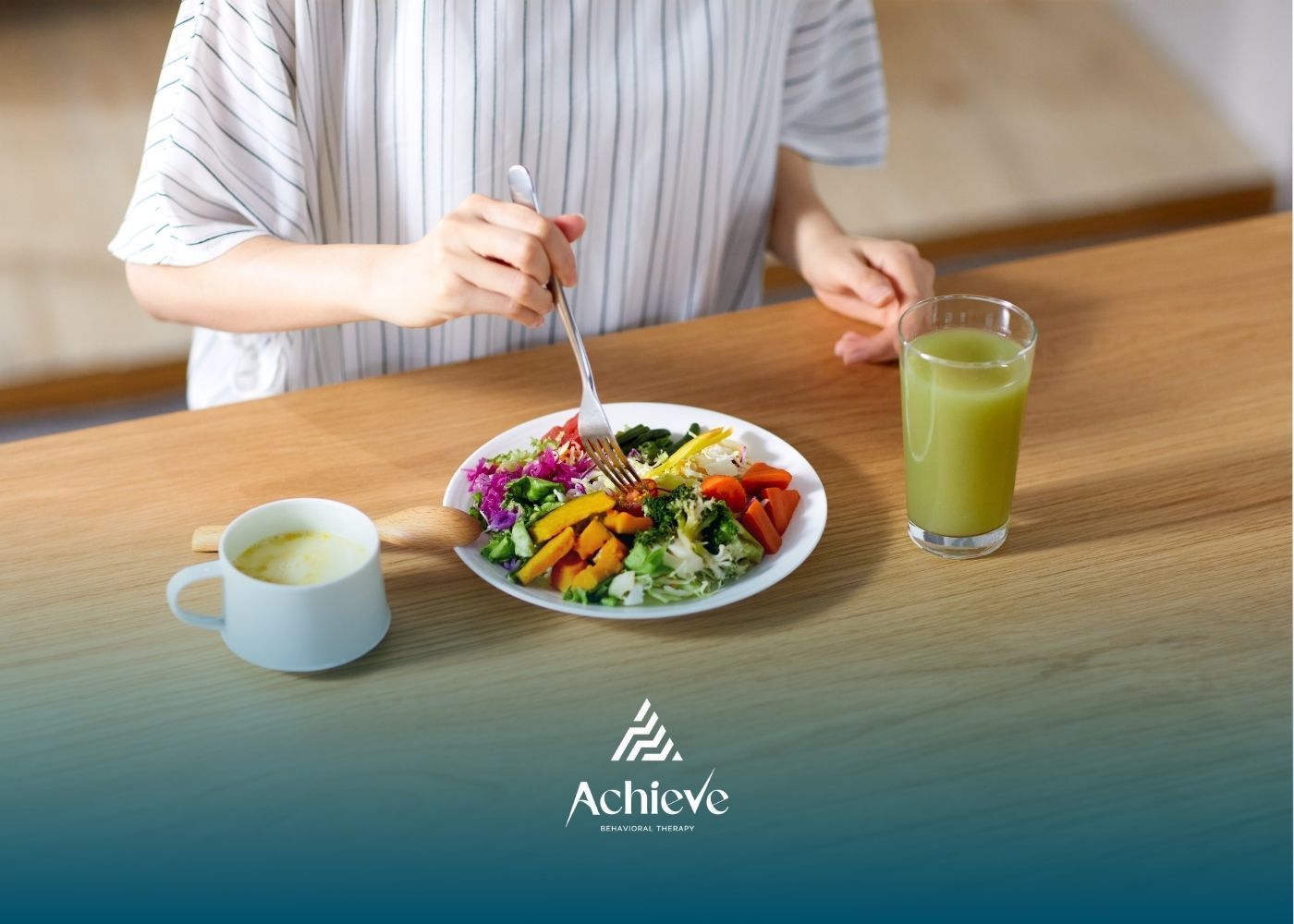 Person eating a colorful salad with a green drink and a mug on a wooden table.