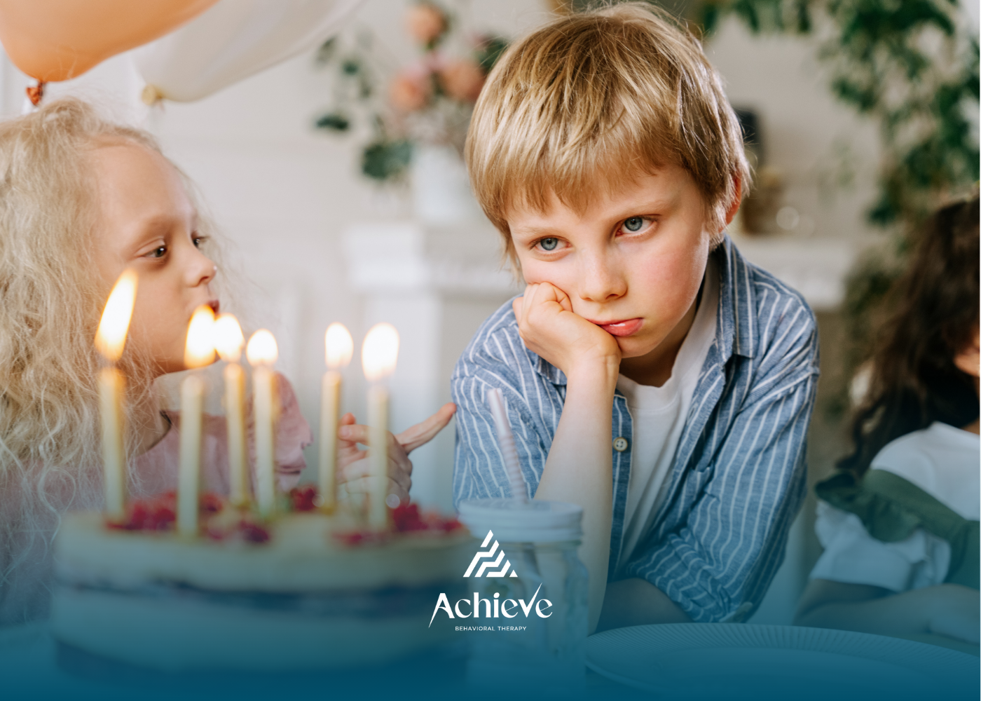 Boy looks unhappy at a birthday party, leaning on his hand. Cake with candles lit in front.