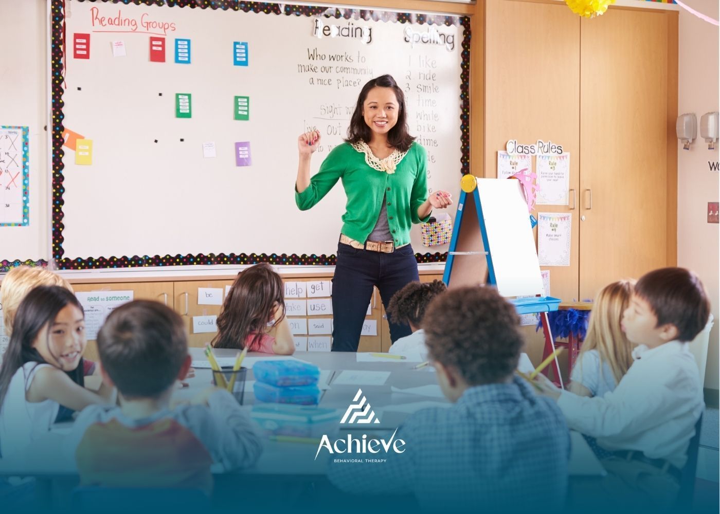 Teacher in green cardigan leading a lesson in a classroom with children sitting at a table.