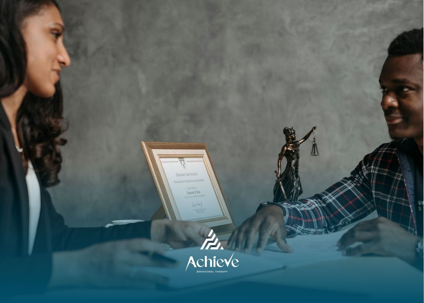 Two people discuss legal matters at a desk with a paralegal certificate and Lady Justice statue