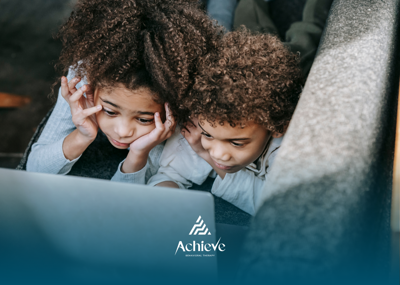 Two young children with curly hair, watching a laptop together, resting their heads on their hands.
