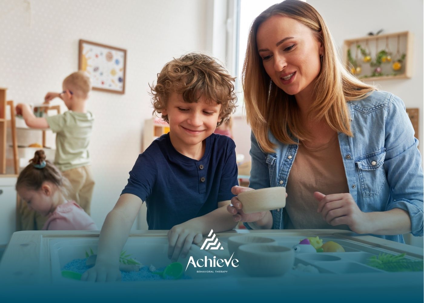 A woman and a young boy interact with a sensory table in a bright classroom with other children playing in the background.