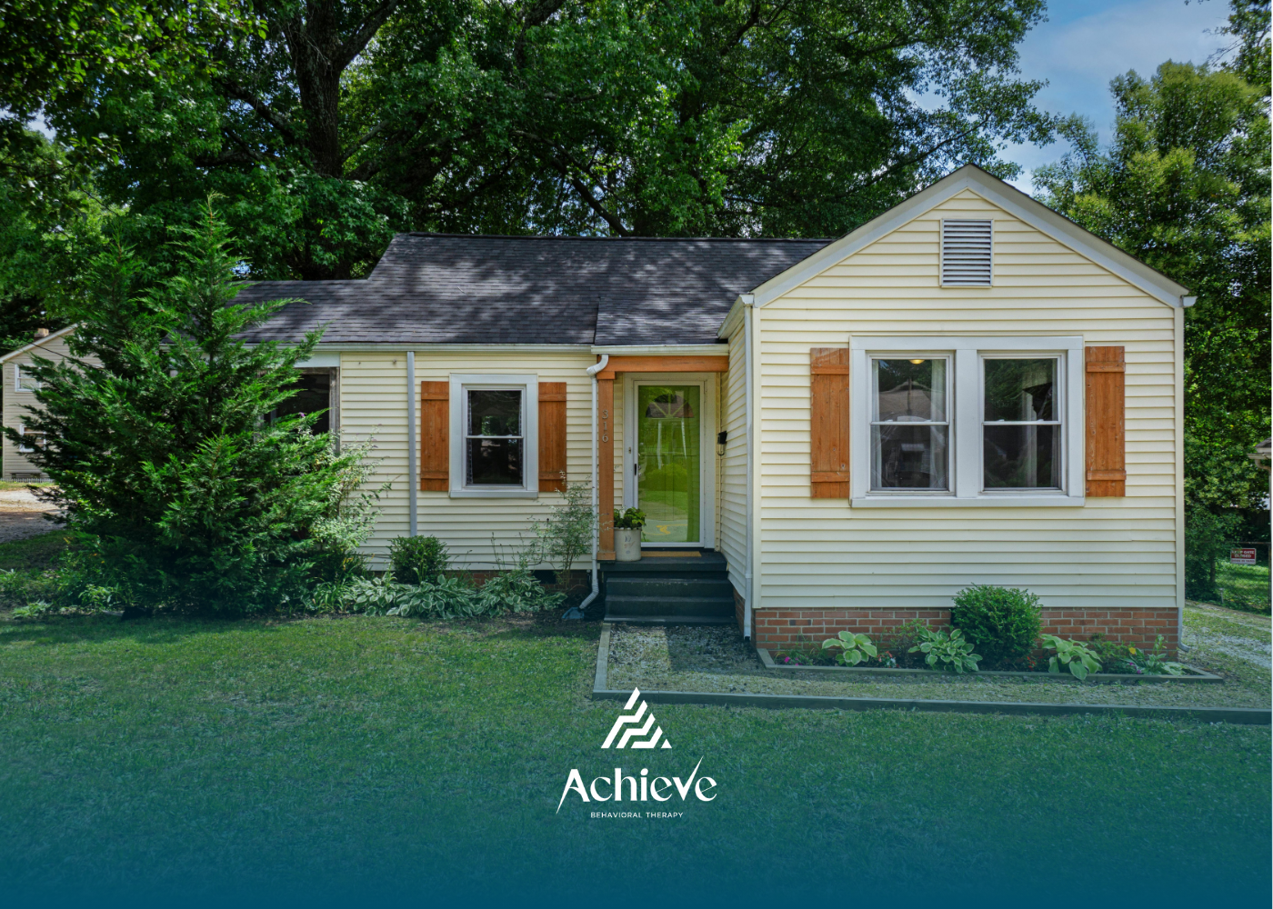 Small, yellow house with brown shutters and green door. Located on green lawn with trees in the background.