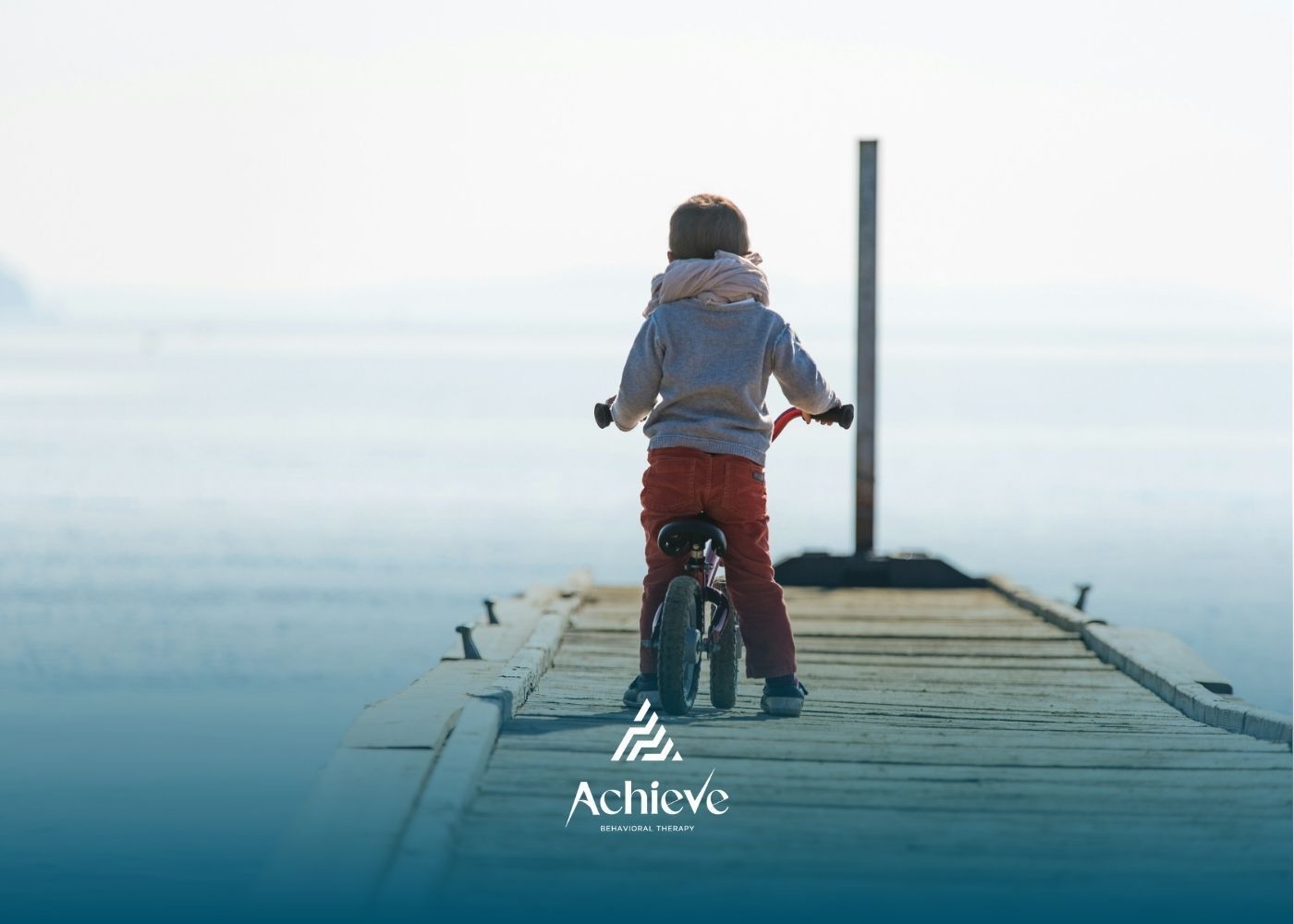 Autistic child rides a small bike on a wooden dock over calm water under soft, hazy lighting.