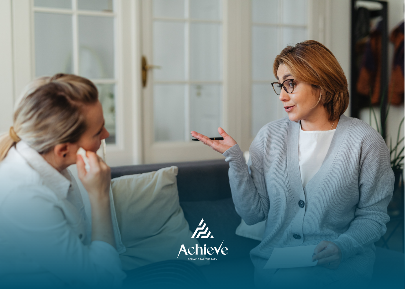 Two people sit on a couch in an office, one holding a tissue and the other gesturing while speaking during a session.