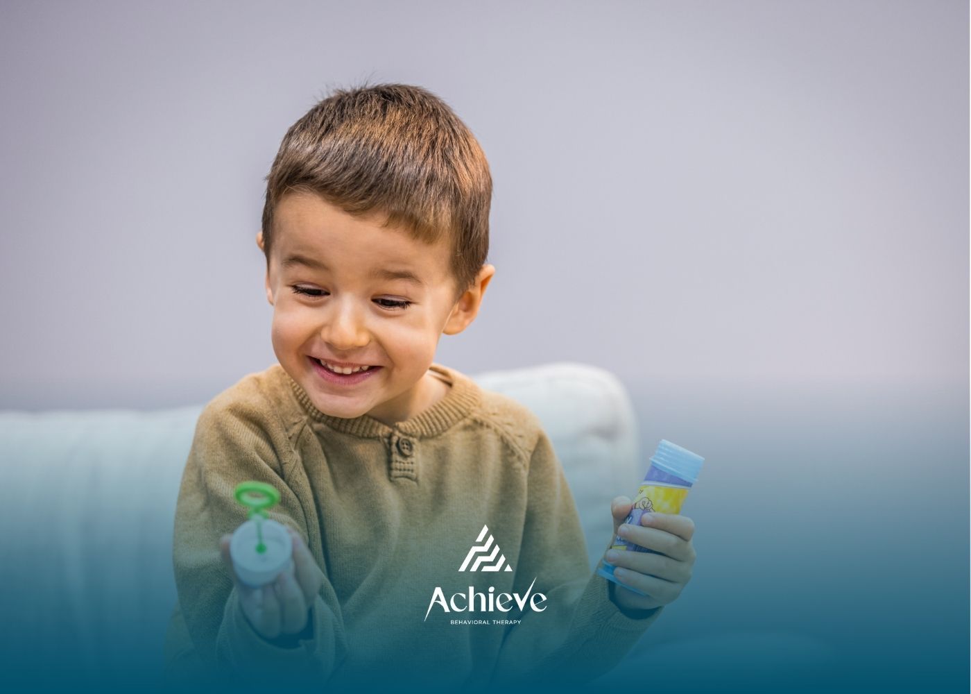 A smiling child plays with bubble wands while sitting on a light blue couch.