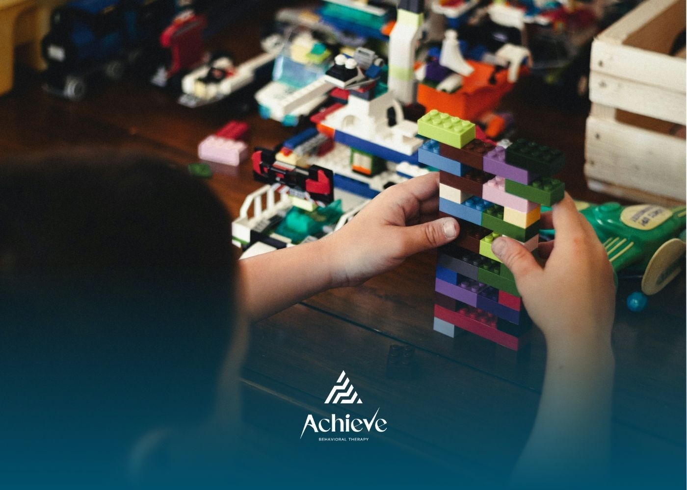 An autistic child builds colorful LEGO tower on wooden surface with toys and models in background.