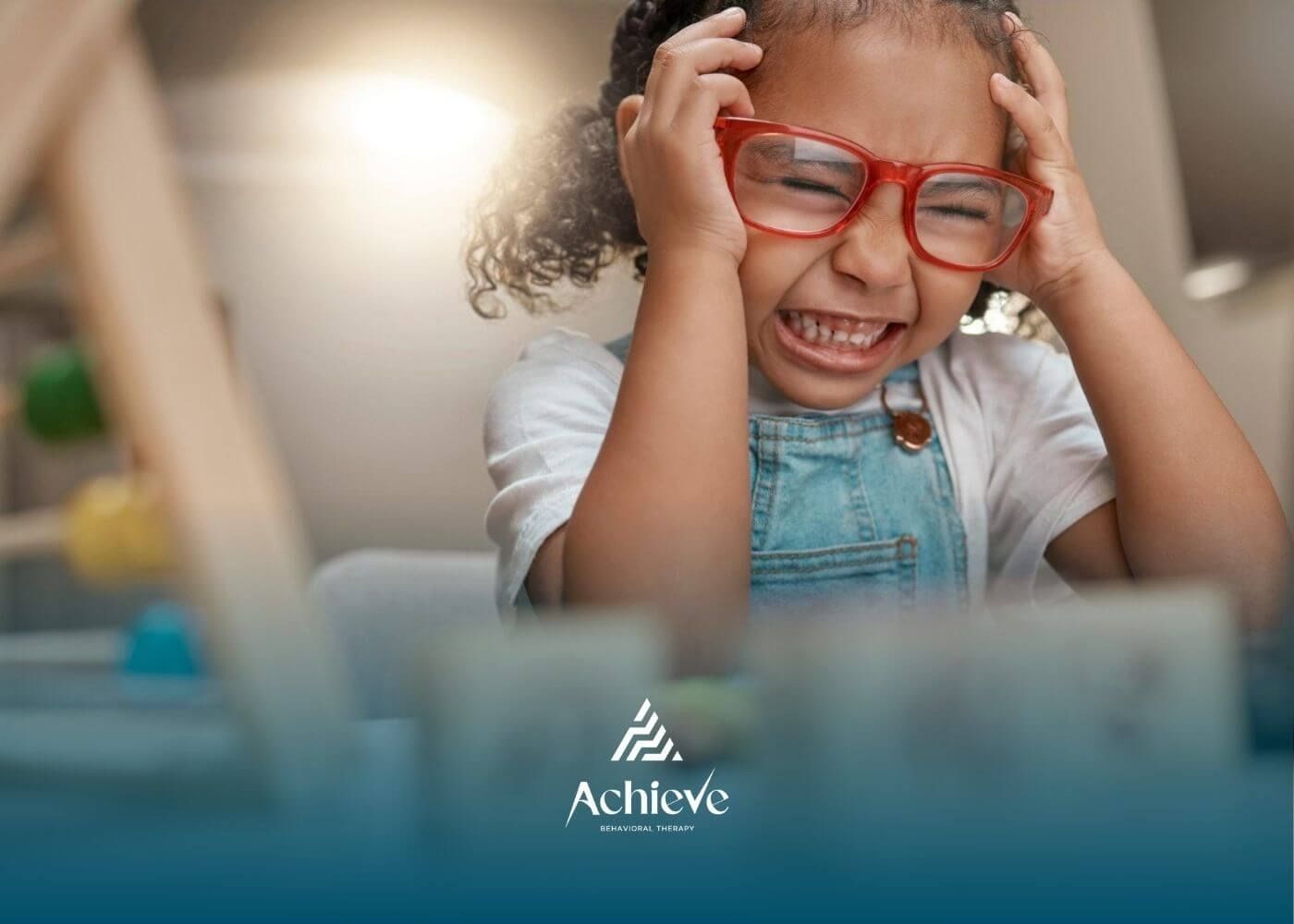 Autistic child with glasses holding head and crying near wooden toys.