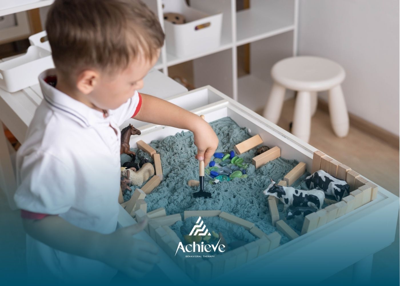 Boy playing with toy farm animals and sand in a sensory bin indoors.